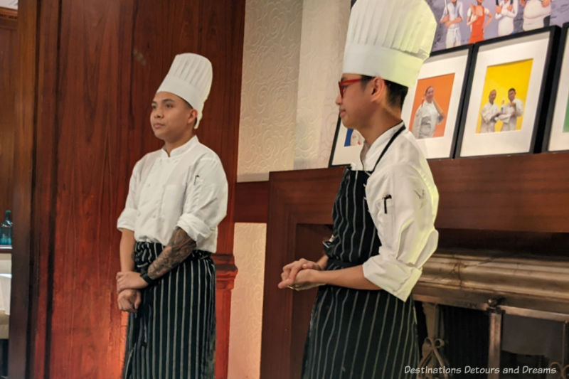 Le Petit Chef Human Chefs Two chefs in white hats, white shirts, and black aprons with white stripes talking to a dining room
