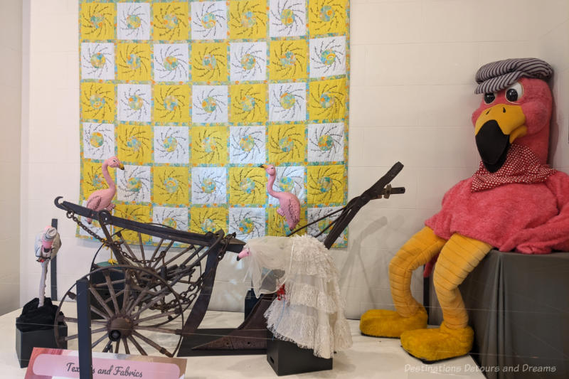 Transcona Museum Textiles Museum display featuring hanging yellow and white quilt, old plough, and stuffed bird
