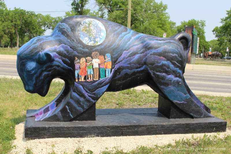 Bison on the Boulevard Only Through Love A black concrete bison with waves of purple and blue, a group of children, and a solar view painted on it
