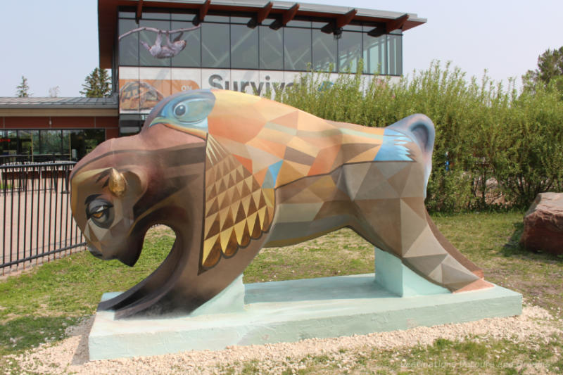 Bison on the Boulevard Piece Together Brown-painted concrete bison containing geometric shapes in yellow, orange and blue hues painted on it as well as a brown cowbird on the back.