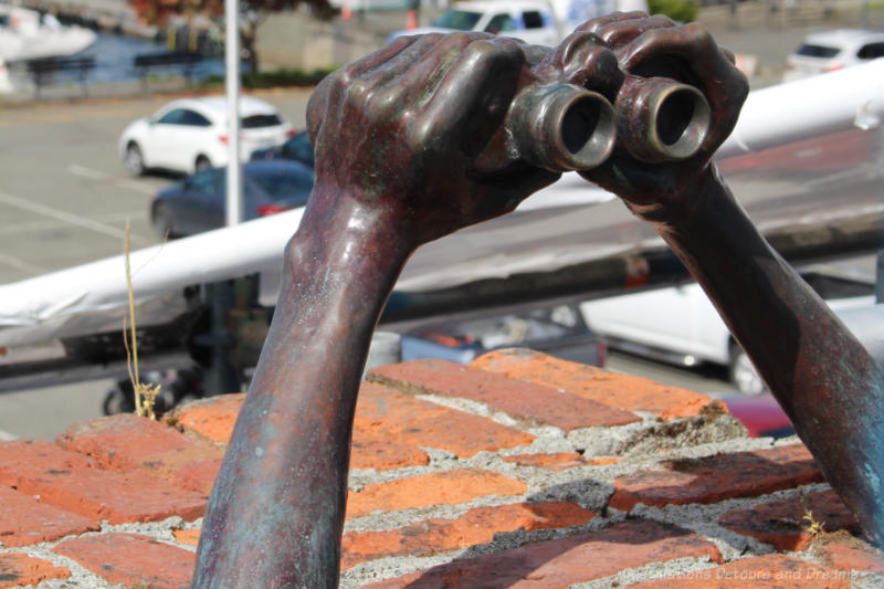 Bronze sculpture resting on top of a red brick wall features arms and hands holding binoculars