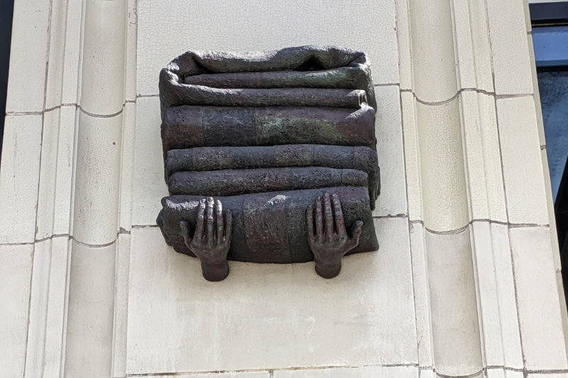 Bronze sculpture of a pair of hands holding a stack of folded blankets on a white brick wall