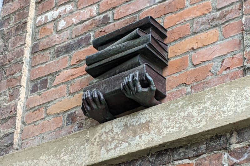 Bronze sculpture on the side of a red brick wall features of a pair of hands holding a stack of books