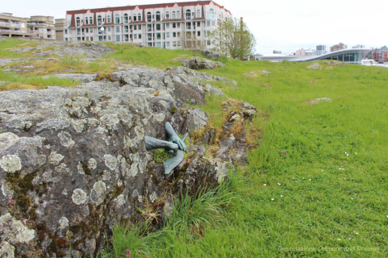Grassy field with a piece of bedrock in foreground which has a bronze sculpture of hands carving a canoe paddle protruding from it