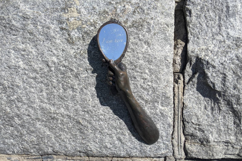Sculpture on the side of a brick wall shows a hand holding a mirror with the words I am here written on the mirror