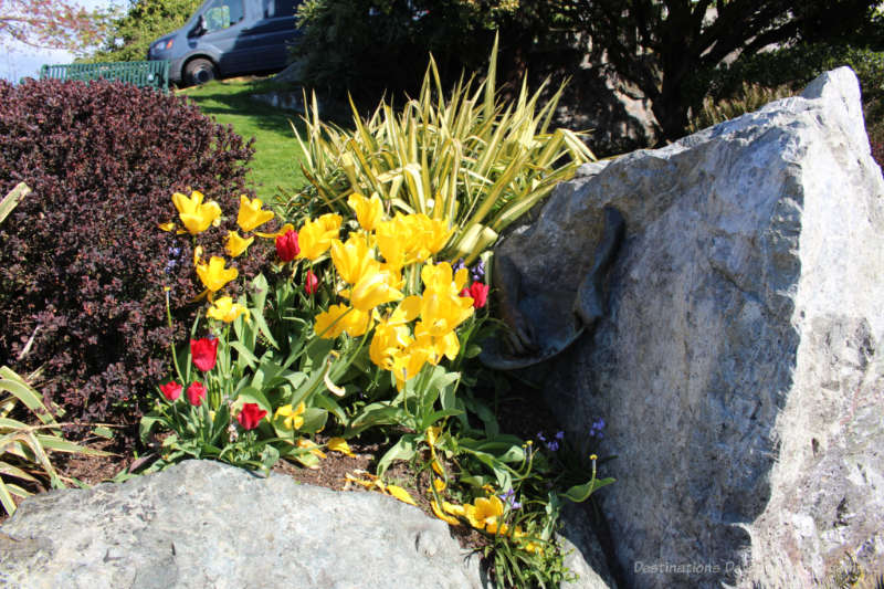 Flower bed with rock in front and to the side and bronze sculpture on the side of the rock at the right