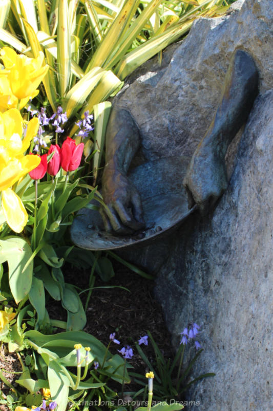 Bronze sculpture on the side of a rock in a flower bed shows hands and a gold panning bowl