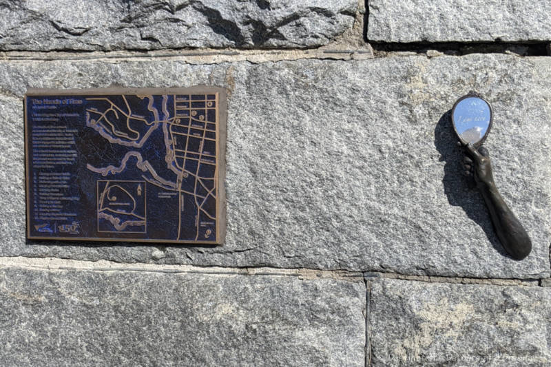 Art installation on a brick wall shows a hand holding a mirror with a bronze map of other installations beside it