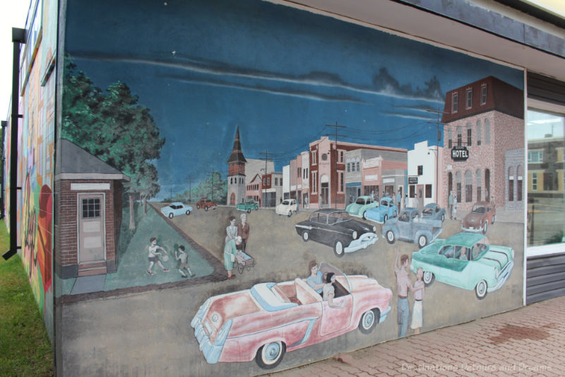 Boissevain Outdoor Art Gallery Main Street Mural on the outside of a building depicts the main street of a small town in the 1950s with cars parker, people walking, hotel at one end, church at the far end