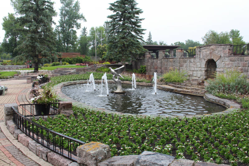 Circular fountain with bird statue in middle, four spouting water fountains around that surrounded by plantings with a brick wall behind it