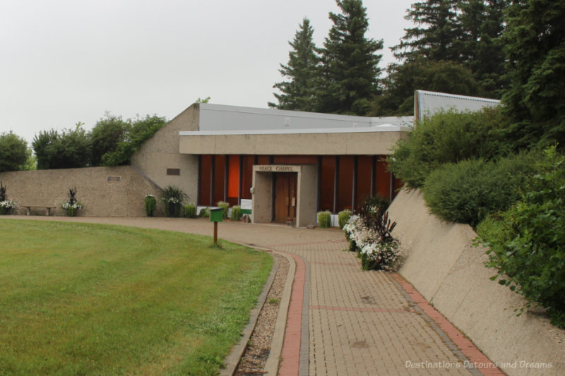 Chapel with wood front with stone walls bordering shrubs along walkway leading to it on either side give the impression of being built into a mountain