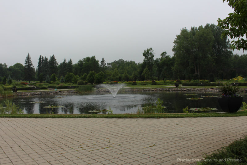 Pond with fountain in middle surrounded by brick walkway and bordered by trees in background