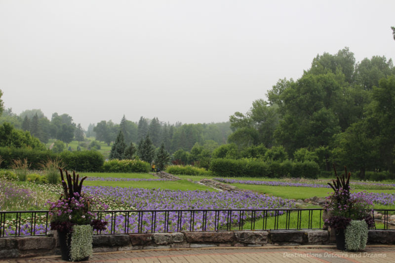 Garden with grassy area intermixed with beds of purple, yellow, and pink flowers and bordered by trees
