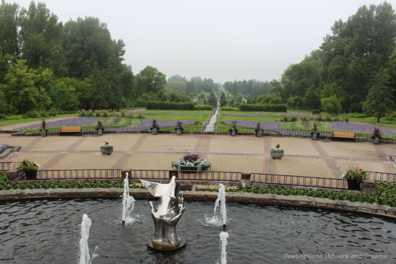 Fountain in front of a brick promenade behind which stretches out a garden with grassy areas, stream now the middle and area with bright purple and other coloured blooms