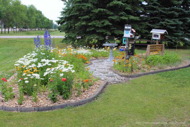 Cement-block bordered garden area on a grassy expanse contain white daisies, blue delphinium, black-eyed susans, red and orange zinnas, a bench a bird bath, a statue of a grain elevator