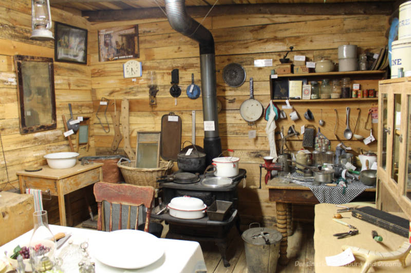 Pioneer kitchen with wood plank walls, cast iron stove, wood shelves and work tables, enamel pots hanging on walls