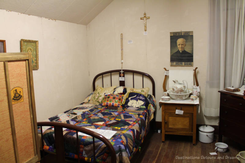Priest room in a museum contain bed with geometric quilt, bedside table with wash basin, crucifix and photo of the priest on wall