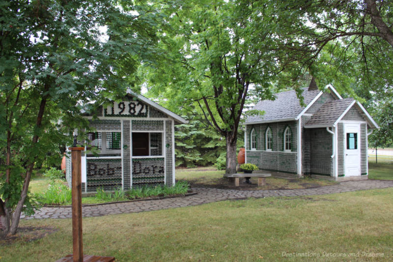 A glass bottle one-room house with the date 1982 and the names Bob and Dora on the front and a glass bottle church in a small park