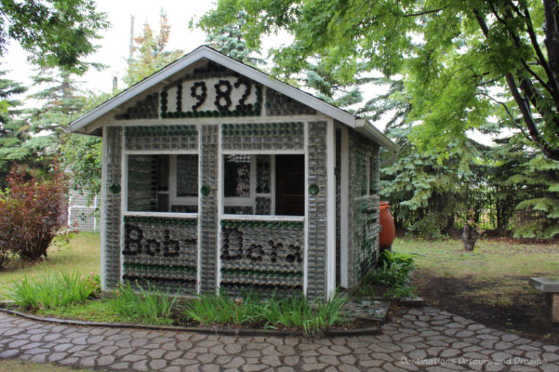 Front of a one-room glass bottle house with pitched roof and the number 1982 and the words Bob and Dora marked on it in bottles