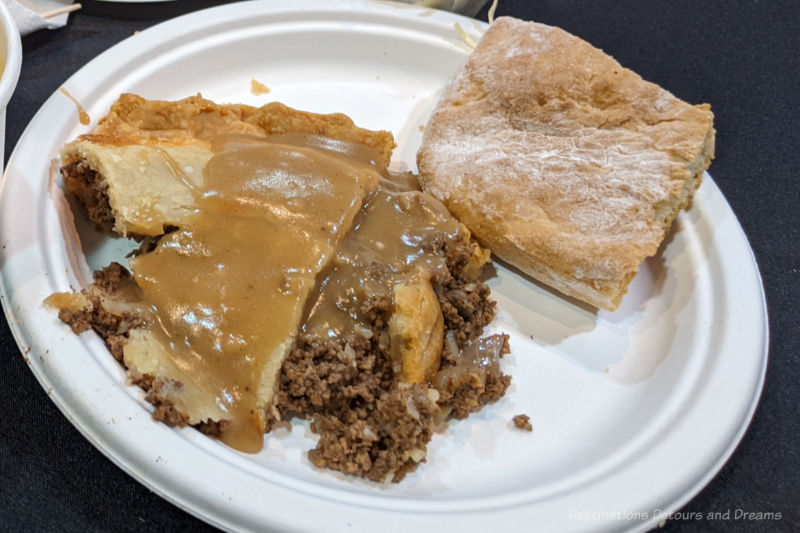 White paper plate containing a slice of tortière with gravy and a piece of bannock
