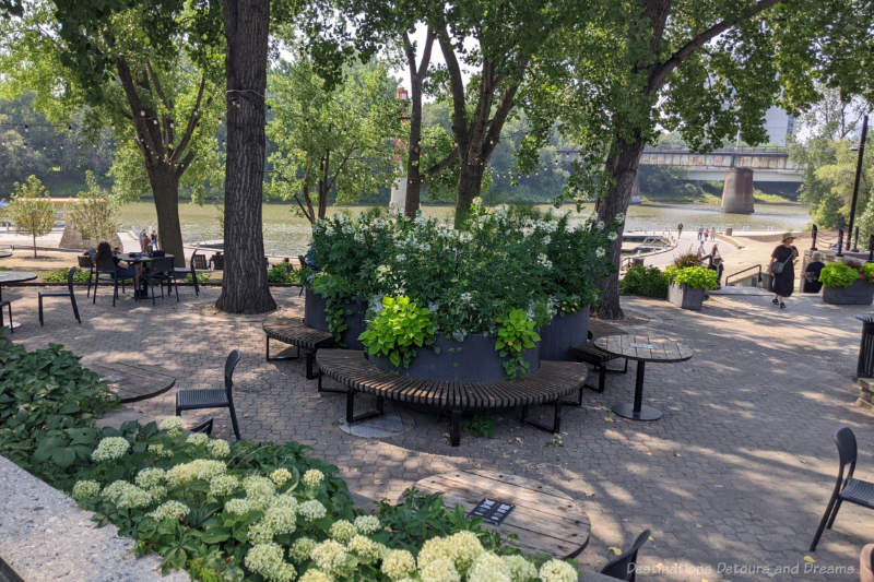 The Forks Turtle Tours Waterfront Stone terraced area along a river waterfront contains trees and shrubs, seating benches and tables