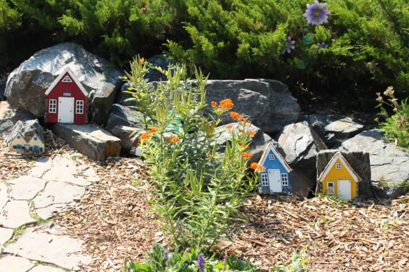 Elf Houses Facade of three tiny fairy houses, one red-brown, one blue, one yellow leaning against rocks in a garden
