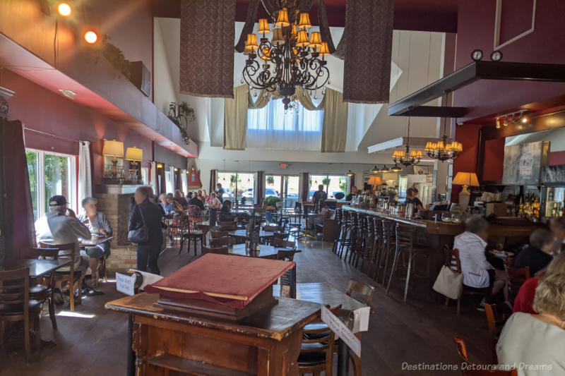 Restaurant dining area with high ceiling, chandelier, wood tables, wooden bar to one side