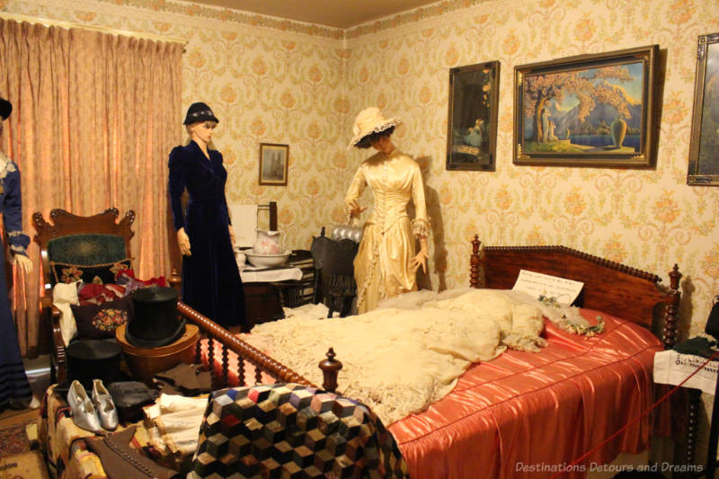 Daly House Museum Master Bedroom Victorian-era bedroom in a house museum features yellow wallpaper, red blanketed bed, washstand, wood chair, and vintage clothing