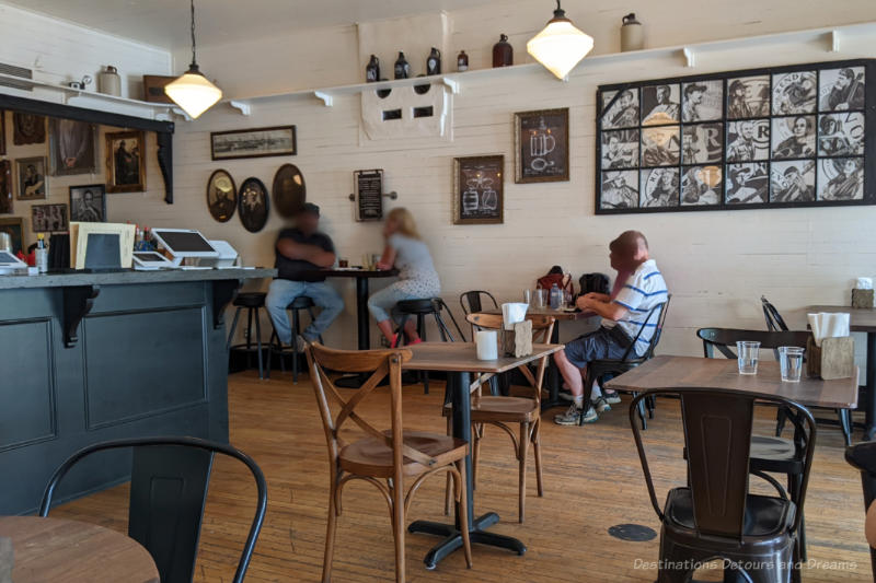 Rendezvous Interior Restaurant with simple tables and chairs, wood floor, white walls with various pictures, and a near ceiling level shelf displaying stoneware kegs