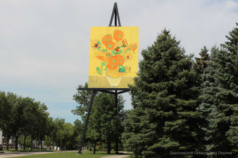 Giant painting of a pot of sunflowers sitting on easel that puts the painting above the trees beside it