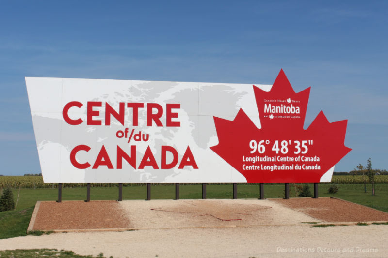 White background billboard announcing in the red lettering the longitudinal centre of Canada and featuring a red maple leaf at one end 