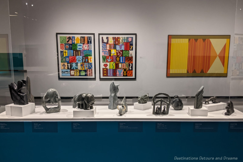 Staycation Collections Display case of sculptures in centre of gallery room with two abstract paintings visible on wall behind