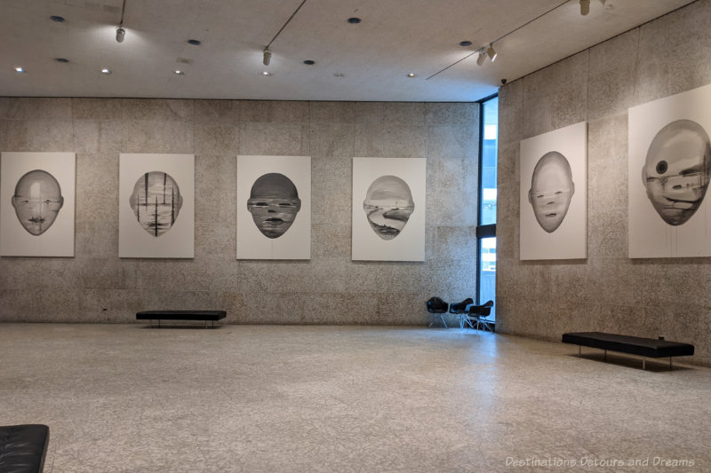 High-ceilinged art gallery space with Tyndall stone floors and walls holding art works featuring immense and surreal human heads