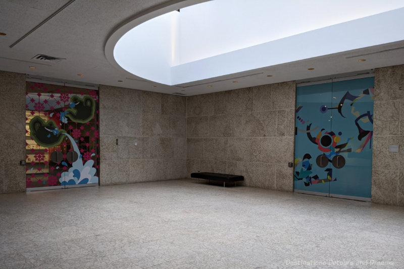 Foyer area with Tyndall stone walls and floor, skylight, and two glass doors covered with brightly coloured collages leading into art gallery space