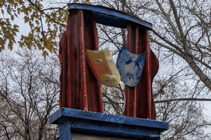 Winnipeg Theatre District Masks Top of a public art sculpture features two theatre masks (one happy face, one sad face) boxed by red metal on the sides and blue on bottom and top