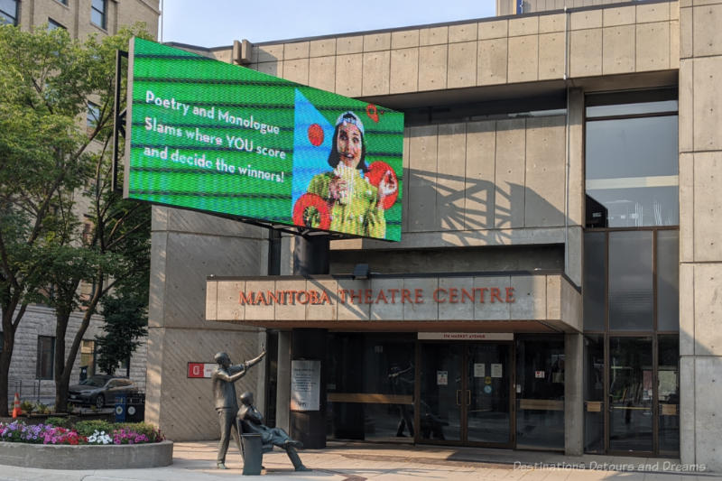 Winnipeg Manitoba Theatre Centre Mainstage Stone front of a theatre centre in Brutalist style architecture with a bronze sculpture in front freatuing one man on park bench facing the theatre and another behind the bench with arms raised toward the theatre