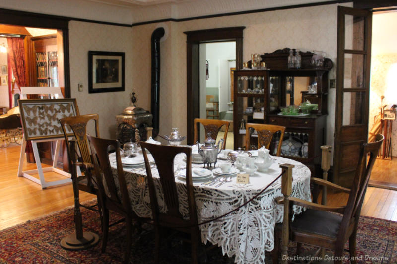 Dining room in house museum decorated as of early twentieth century with dark wood chair, dining hutch, and table covered with white lace tablecloth, patterned wallpaper and dark wood door moldings.