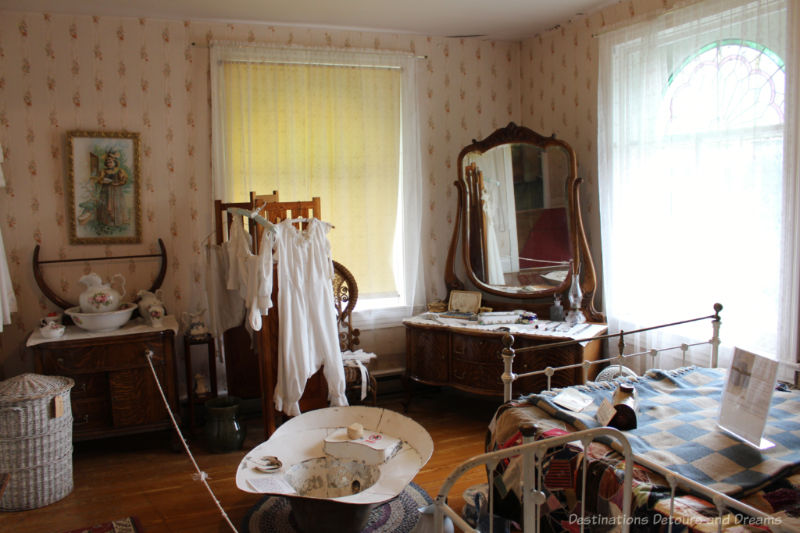 Bedroom ala early twentieth century in house museum feature white metal-frame bed, wash basin table, dresser with mirror in curved frame, clothing hanging on divider, and metal wash tub
