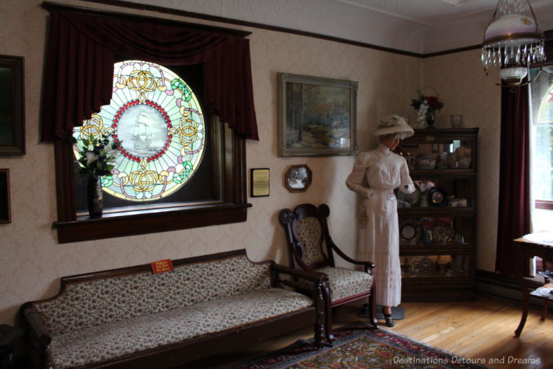 House museum room of early twentieth-century home featuring plank floors, circular stained glass windows, patterned wallpaper, ornate ceiling, period settee, chair, and china cabinet