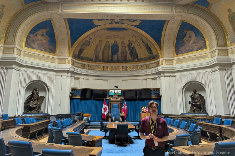 Legislative Assembly Room with blue carpet and blue chairs around wood tables arranged in a horseshoe and with murals painted on upper walls