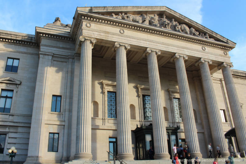 Entrance to a several story stone legislative building fronted by ionic columns atop which sits a triangular pediment with carved figures