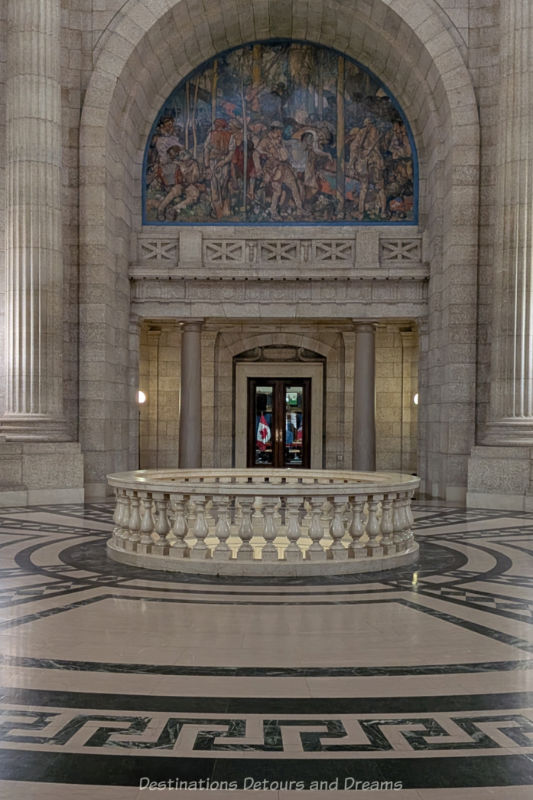 Rotunda area in legislative building with stone walls, marble floors in continuous Greek key design, a round marble railing in centre around hole looking to floor below, and a large war memorial mural above the door to the Legislative Assembly