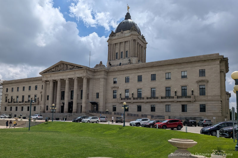 The front of a 1920 stone legislative building with decorative features, columns in front of entry, and a central dome topped with a golden statue of a young man