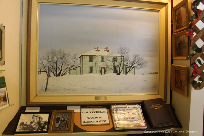 Sipiweske Museum Criddle and Vane Legacy Museum display showing painting of Criddle and Vane homestead with collection of photos and books on a shelf below it
