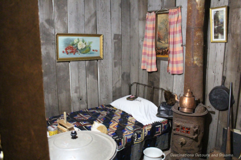 Sipiweske Museum Pioneer Life Sleeping Museum exhibit of pioneer life showing sleeping cot and wood-burning heating stove in room with wood-plank walls