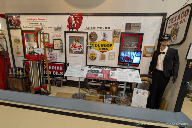 Museum display features vintage garage signs, a mannequin in a chauffer outfit, hubcaps, and oil cans