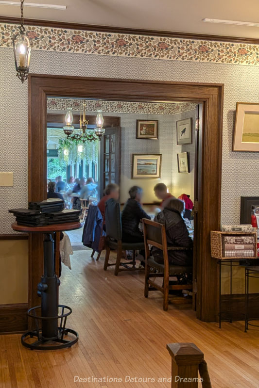Wide wood-framed doorway in a wall-papered wall of an old house leads to a small room and through that to a closed-in veranda with tea room dining tables