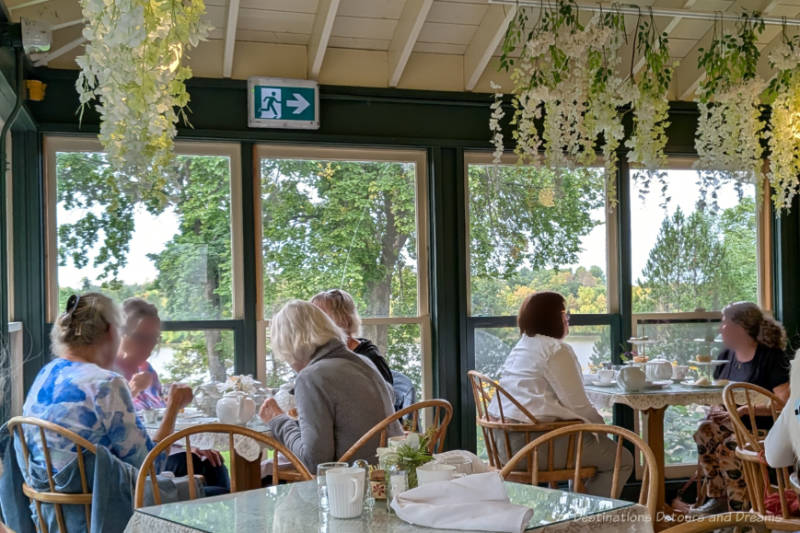 Tea room in a closed-in veranda with walls of windows looking out over trees and riverbank and containing tables with glass-topped white tablecloths