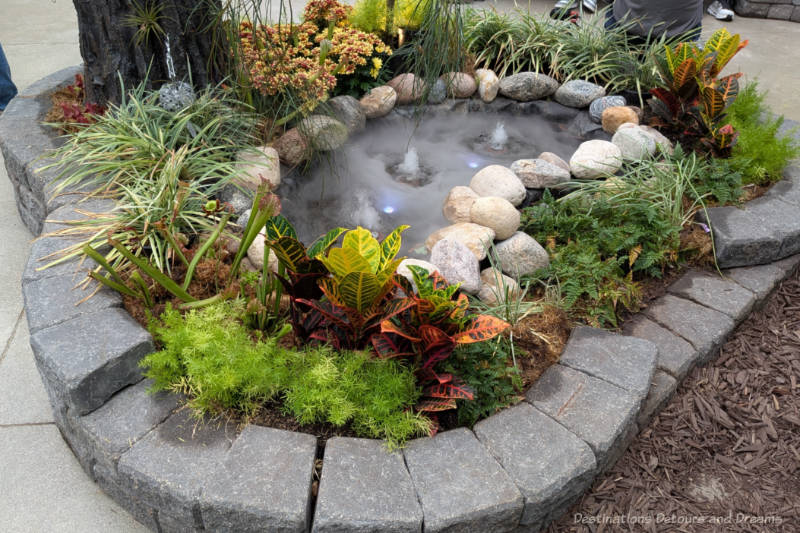 Pond surrounded by stones and foliage has three small waterfalls generating fog