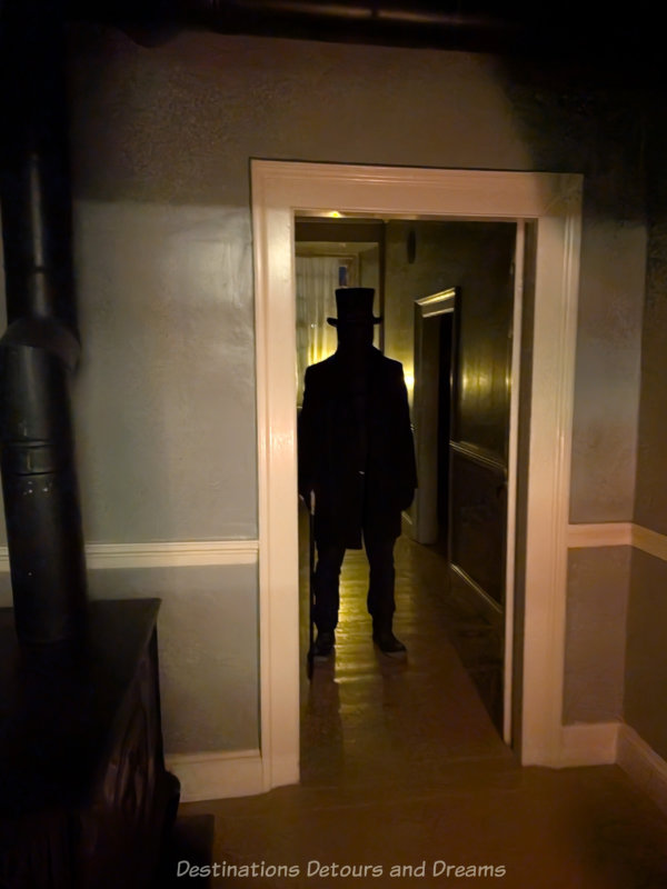 Figure dressed all in black and wearing a black top hot standing behind doorway of dimly lit house
